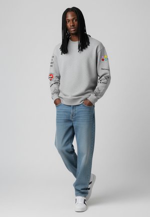 Felpa grigia con dettagli grafici colorati sulle maniche, abbinata a jeans azzurri chiari e sneaker bianche con accenti neri.
