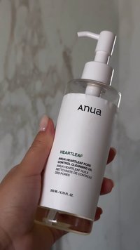 Mano che tiene una bottiglia trasparente di Anua Heartleaf Pore Control Cleansing Oil, 200 ml, su uno sfondo di marmo.