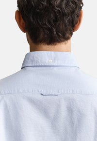Mann mit kurzen lockigen dunklen Haaren, der ein hellblaues Hemd mit Knöpfen trägt, von hinten vor einem einfarbigen Hintergrund gezeigt.