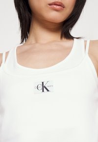 Biały prążkowany top na ramiączkach z cienkimi paskami, z prostokątną łatą z logo "Calvin Klein Jeans" w czerni i szarości.