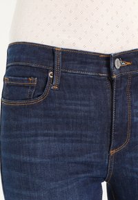 Jeans de mezclilla azul oscuro con una textura suave. Presentan costuras naranjas, un bolsillo delantero y un cierre de botón de metal en la cinturilla.