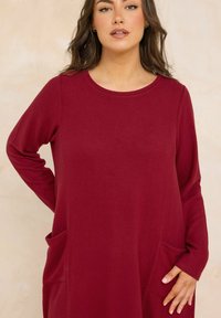 Vrouw met golvend bruin haar, gekleed in een maroon lange mouwen jurk met zakken, staande tegen een beige achtergrond.