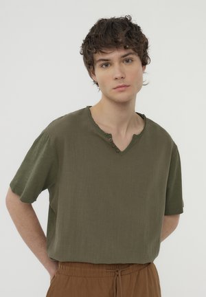Camicia verde oliva a maniche corte con scollatura a V e tre bottoni decorativi sul collo. Tessuto morbido e texturizzato con una vestibilità ampia.