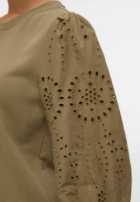Camisa caqui con mangas abullonadas que presenta intrincados patrones florales recortados y bordados. Tejido de algodón suave con detalle texturizado.