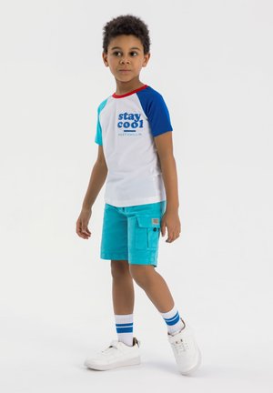 Lyhythihaisella valkoisella T-paidalla, jossa on sinisiä ja punaisia yksityiskohtia, ja jossa lukee "stay cool". Yhdistetty turkoosien cargo-shortsien ja valkoisten lenkkareiden kanssa.