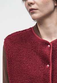 Žena koja nosi teksturirani smešani prsluk boje tamnocrvene s dugmadima preko rebrastog smešano smešane boje, prikazana od vrata do ramena.