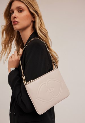 Borsa a tracolla in pelle rosa chiaro con design a cuore in rilievo, tracolla in metallo dorato e chiusura a zip, indossata sulla spalla con un blazer nero.