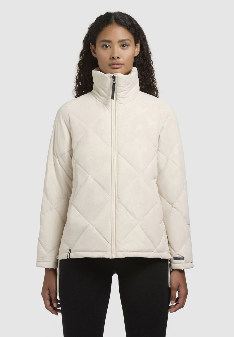 Chaqueta acolchada y puf, de color crema, con un alto cuello, que cuenta con un cierre frontal completo y bolsillos laterales con cremallera. Textura suave con patrón de diamantes.