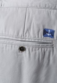 Světle šedá látka s hladkou texturou, která má přišitou kapsu s modrou nálepkou zobrazující bílý design kotvy a kulatý černý knoflík.