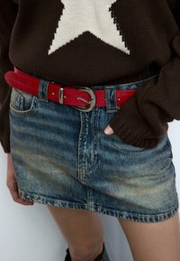 Minigonna in denim con lavaggio blu sbiadito, abbinata a una cintura in pelle rossa a motivo coccodrillo e a un maglione marrone con una stella bianca.
