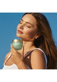 Hydratant léger dans un pot vert translucide avec un couvercle doré. Modèle tenant le pot contre un ciel bleu clair, portant un haut blanc à volants.