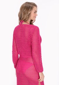 Copertura in crochet rosa con maniche lunghe e design a rete aperta. Presenta un nastro in vita e una texture leggermente trasparente per una sensazione di leggerezza.