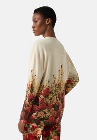 Kremowy kardigan z kwiatowym nadrukiem w odcieniach czerwieni, różu i zieleni. Charakteryzuje się luźnym krojem, ściągaczowymi mankietami oraz miękką fakturą.