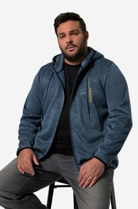 Felpa con zip di colore blu navy in un tessuto testurizzato, con tasche frontali e rifiniture nere a contrasto, abbinata a jeans grigi e una maglietta nera.