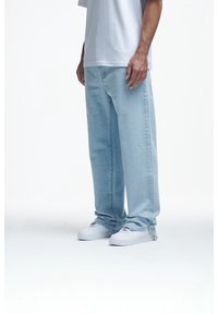Hombre vistiendo jeans de pierna ancha de color azul claro, zapatillas blancas y una camiseta blanca, de pie contra un fondo blanco liso.