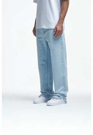 Homme portant un jean large bleu clair, des baskets blanches et un t-shirt blanc, se tenant devant un fond blanc uni.