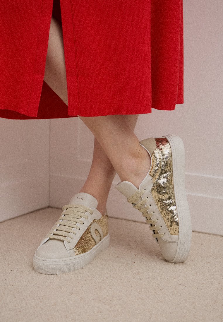 Pies cruzados con zapatillas de lentejuelas beige y doradas, combinadas con una falda roja, de pie sobre una alfombra clara cerca de la esquina de una pared blanca.