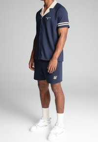 Navy blue short-sleeve button-up shirt with cream collar and stripes, paired with matching shorts featuring embroidered logo, worn with white sneakers.