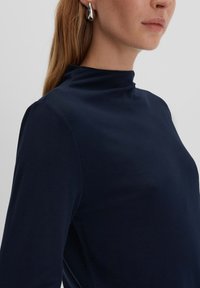 Navyblaues Langarmoberteil mit hohem Ausschnitt, glatter Textur und taillierter Passform, kombiniert mit einem markanten silbernen Ohrring.
