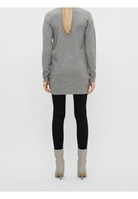 Woman with long blonde hair wearing a gray long-sleeve sweater, black leggings, and light gray high-heeled ankle boots, standing facing away.