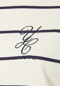 Vit randig skjorta med marinblå horisontella ränder. Broderad logotyp i marinblått som säger "GC LONDON" under logotypen. Klassisk rundhalsad design.