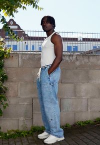 Világoskék farmer baggy nadrág fehér Nike logóval, fitted fehér ujjatlan toppal és fehér sportcipővel párosítva; egy szürke fal előtt áll.