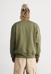 Oliivinvihreä collegepaita resorilla kaula-aukon ja hihansuiden kohdalla, valmistettu sileästä puuvillasekoitteesta. Päällä on beiget cargo-housut, takaapäin nähtynä.