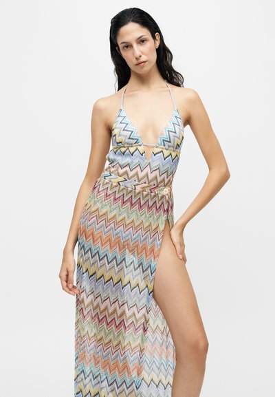Γυναίκα με πολύχρωμο φόρεμα halter neck με ζιγκ ζαγκ μοτίβο και ψηλή σχισμή, ποζάρει με το ένα χέρι στη μέση μπροστά από απλό φόντο.