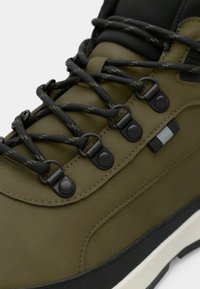 Olivengrønn joggesko med teksturert syntetisk overdel, svarte lisser, metallfester og en kontrasterende hvit såle.