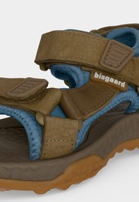 Sandale à bout ouvert avec des brides marron réglables, une doublure matelassée bleue, une semelle texturée et une étiquette "bisgaard" sur la bride latérale.