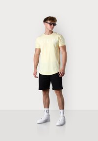 Yellow short-sleeve t-shirt with white text, paired with black shorts, white sneakers, and sunglasses. Smooth fabric texture.