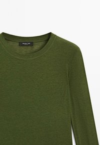 Camicia a maniche lunghe in tessuto verde scuro con scollatura tonda. Presenta una texture liscia e dettagli di cucitura minimi. Etichetta visibile.