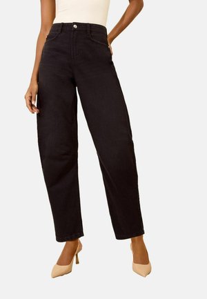 Vrouw draagt zwarte high-waisted jeans met wijde pijpen en beige hoge hakken tegen een effen achtergrond.