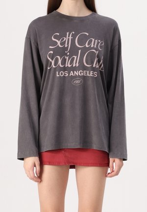 Person trägt ein graues Langarmshirt mit der Aufschrift "Self Care Social Club Los Angeles 1995" und einen roten Minirock.