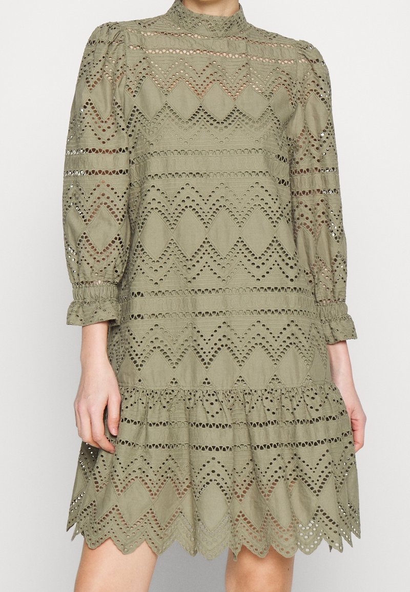 Robe vert olive avec un col montant, des manches bouffantes longues et des découpes présentant des motifs en zigzag et un ourlet froncé.