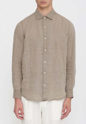 Uomo che indossa una camicia marrone chiaro a maniche lunghe con bottoni e colletto e pantaloni bianchi, in piedi contro uno sfondo semplice.