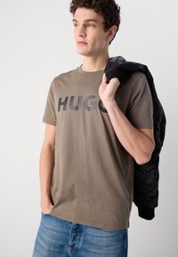 Brūns kokvilnas T-krekls ar "HUGO" logo tumši pelēkā krāsā. Modelis valkā zilos džinsus, tur rokā melnu bomber jakku. Vienkāršs apaļas kakla dizains.