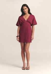 Vestido corto burdeos con escote en V profundo, mangas abullonadas y detalle fruncido en la cintura. Presenta un diseño simple y ajustado, con una textura suave.