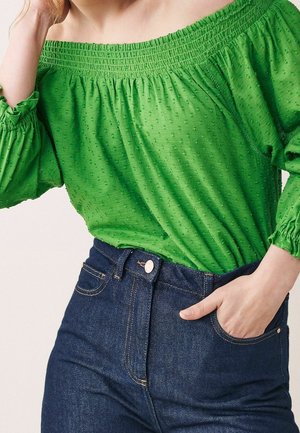 Vrouw draagt een groene off-shoulder textuur blouse met elastische halslijn en mouwen, gecombineerd met een donkerblauwe high-waisted jeans, hand in de zak.