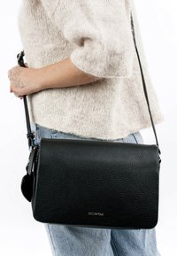 Bolso cruzado de piel negra con acabado texturizado, forma rectangular y correa ajustable. Presenta un sutil acento de hardware.