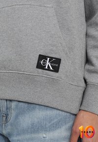 Šedá mikina s žebrovanými manžetami a lemem, která má přední kapsu a černý štítek s logem "CK" a značkou "Calvin Jeans".