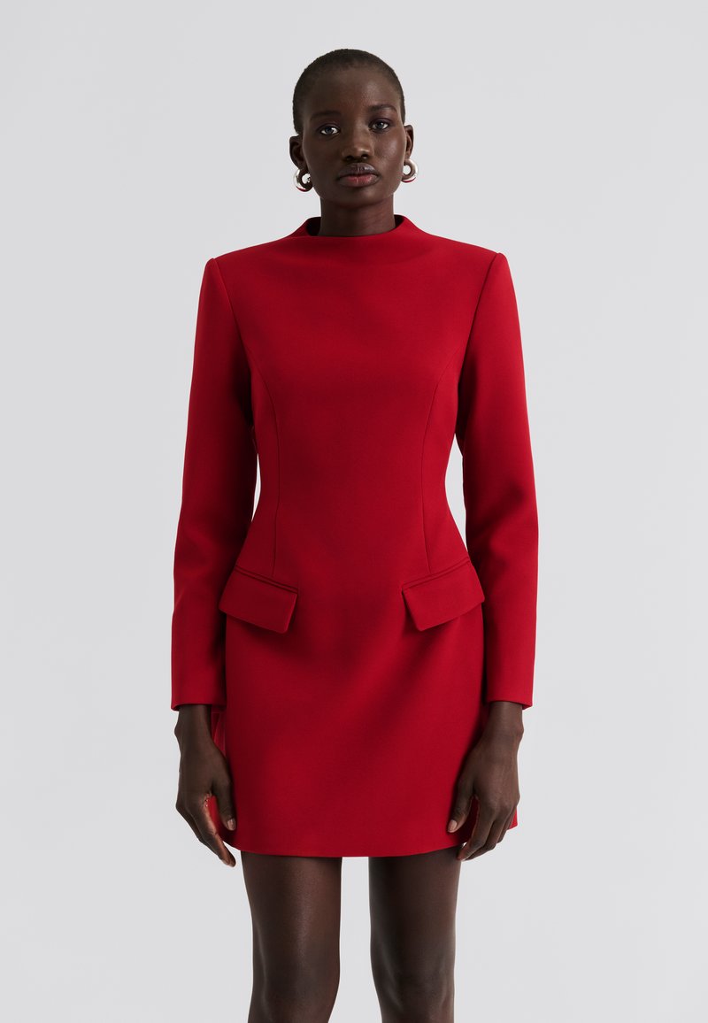 Piros ruha hosszú ujjakkal, magas nyakkal és strukturált vállakkal. Két első zsebbel és testhez simuló, letisztult vonalvezetésű sziluettel rendelkezik.