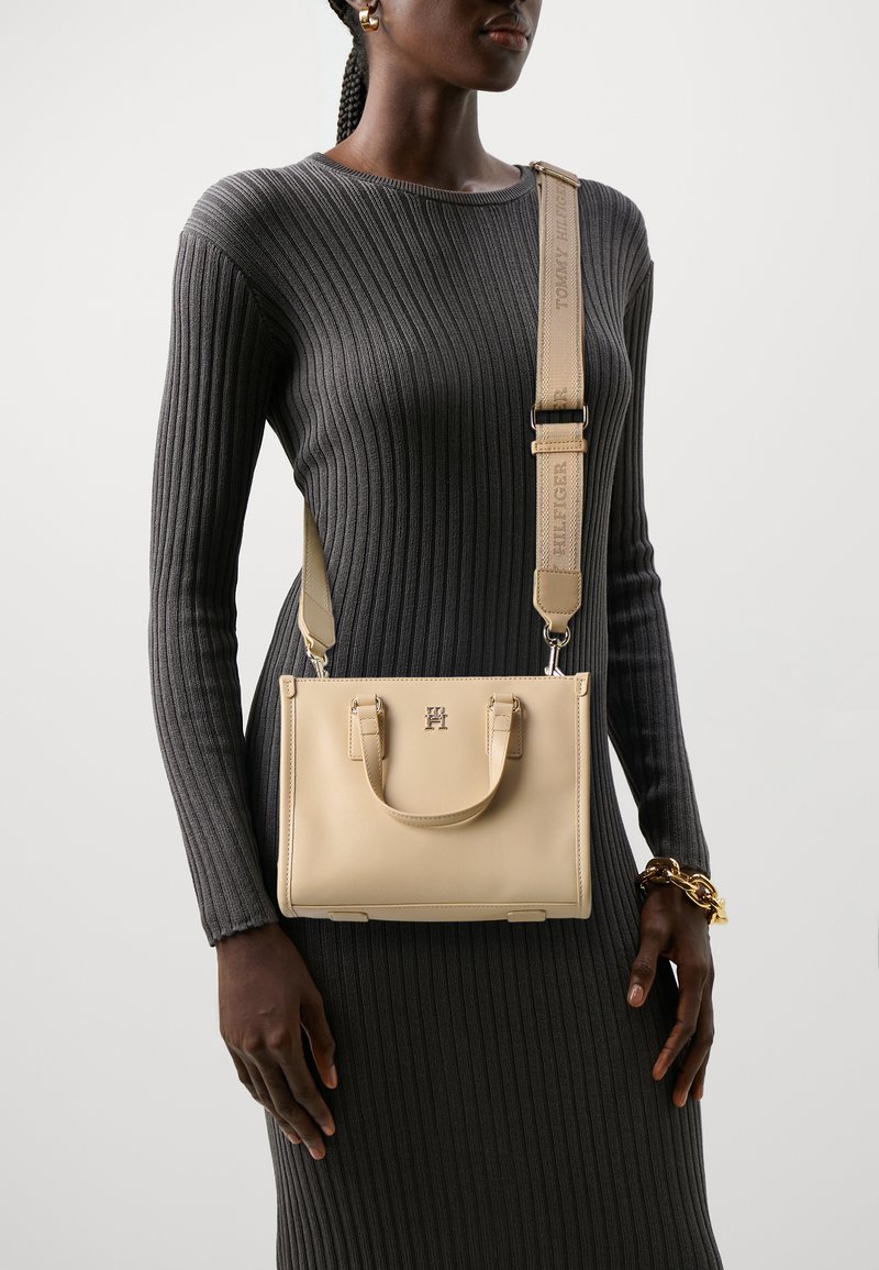 Lys beige håndveske med en strukturert form, topphåndtak og en avtakbar skulderrem med "TOMMY HILFIGER"-merke. Glatt tekstur.