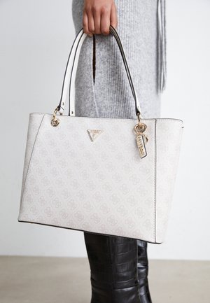 Main tenant un sac cabas blanc à motifs Guess avec un pendentif logo doré, porté avec une robe en maille grise et des bottes noires texturées.