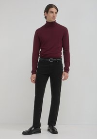 Uomo con un dolcevita borgogna, cintura nera, pantaloni slim-fit neri e mocassini in pelle nera, in piedi contro un muro chiaro e senza decorazioni.