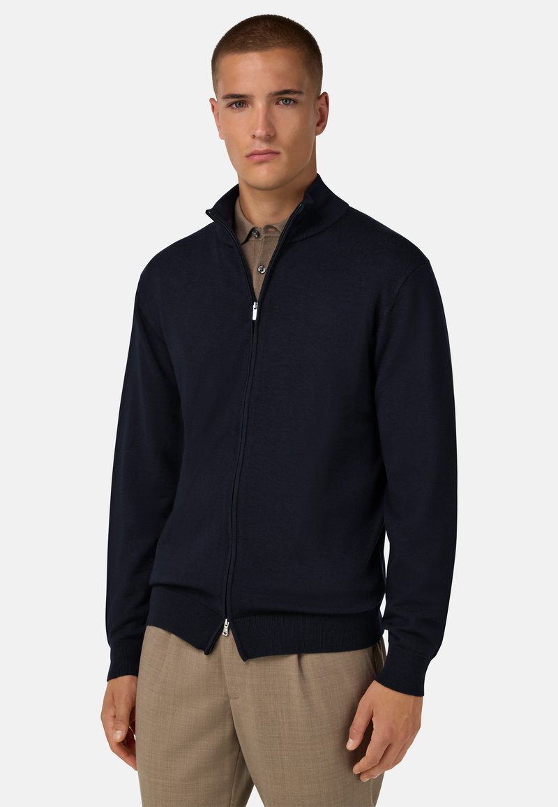 Jūrinės jūrų tamsiai mėlynos spalvos užtrauktukas megztinis su aukštu apykakle, raukšlėtomis rankovėmis ir apačia, pagamintas iš lygios medžiagos, uždėtas ant smėlio spalvos marškinėlių.