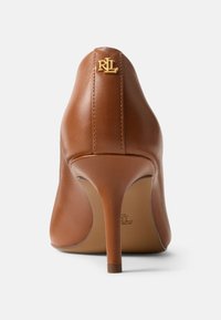 Hnědá kožená lodička na vysokém podpatku s špičatou špičkou, zdobená zlatým logem na zadní straně a tenkým jehlovým podpatkem. Hladká textura.