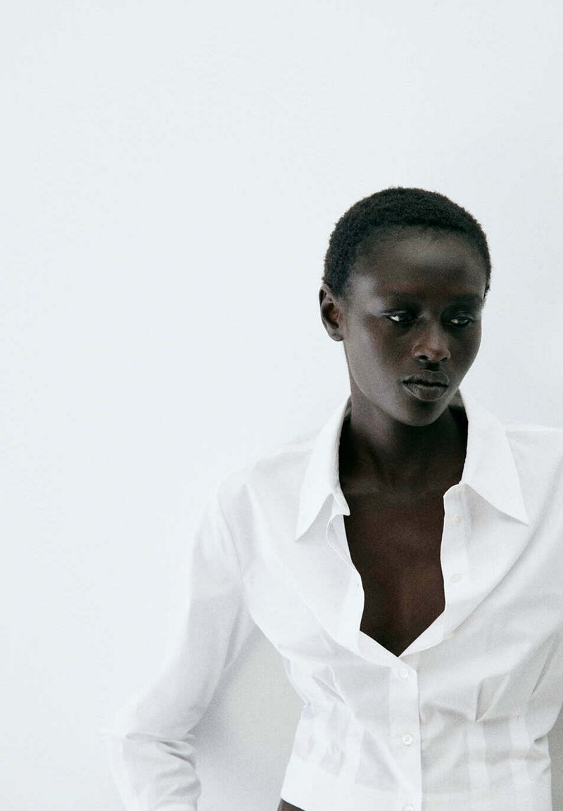 Femme aux cheveux courts portant une chemise blanche ouverte à boutons, regardant vers le bas et légèrement sur le côté sur un fond blanc uni.