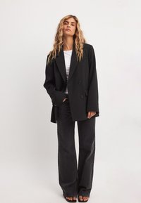 Musta kaksirivinen bleiseri ylipitkässä mallissa, jossa on olkatoppaukset ja sivutaskut, yhdistettynä tummiin leveälahkeisiin farkkuihin ja korkokenkiin.