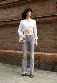 Blusa branca de mangas longas com detalhe de nó, jeans cinzentos de cintura alta, sandálias pretas de slide, mala transparente, contra uma parede de tijolos.
